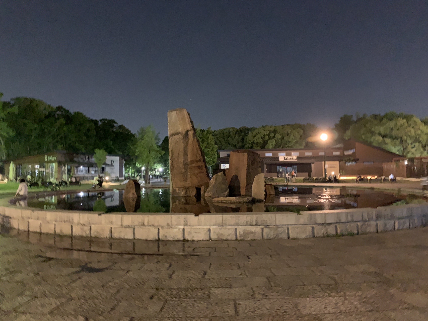 公園内の噴水と石のモニュメント。奥に建物の明かりが灯っている