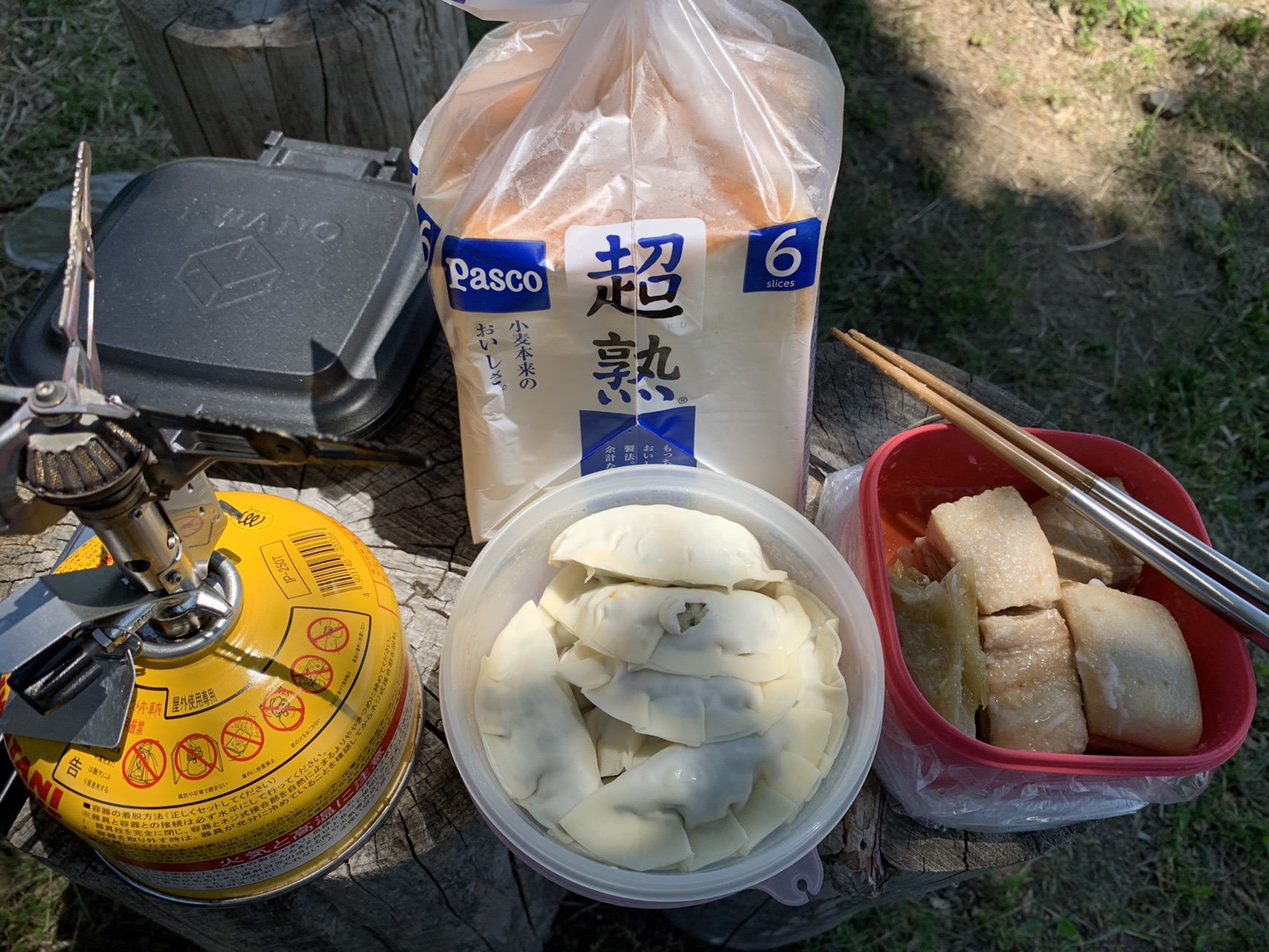 切り株の上に並んだホットサンドメーカー、食パン、タッパーに入った餃子と角煮