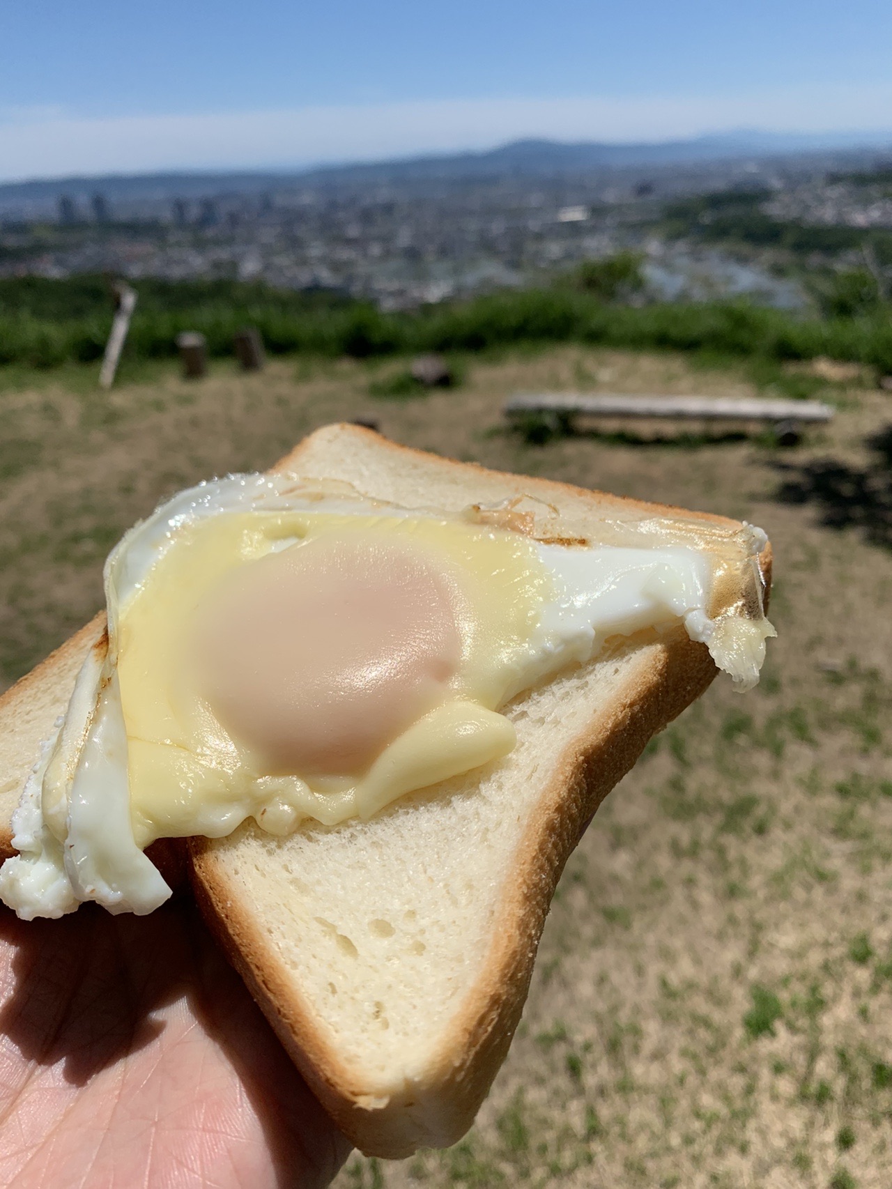 食パンの上に目玉焼きとチーズがのった、手に持たれたトースト。奥に街の眺め