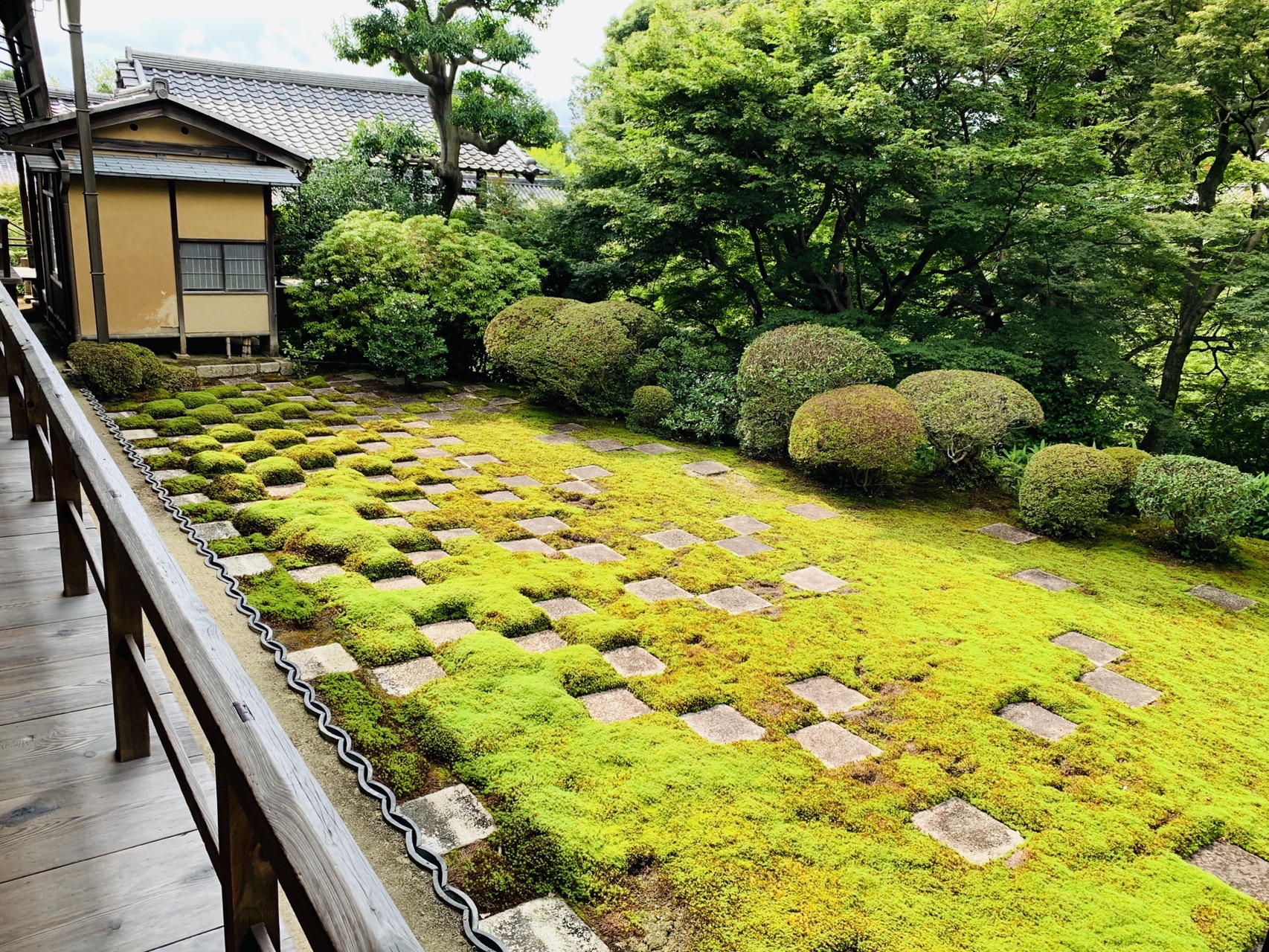 苔の中に四角い切石が市松模様に並ぶ庭。奥に建物と木立
