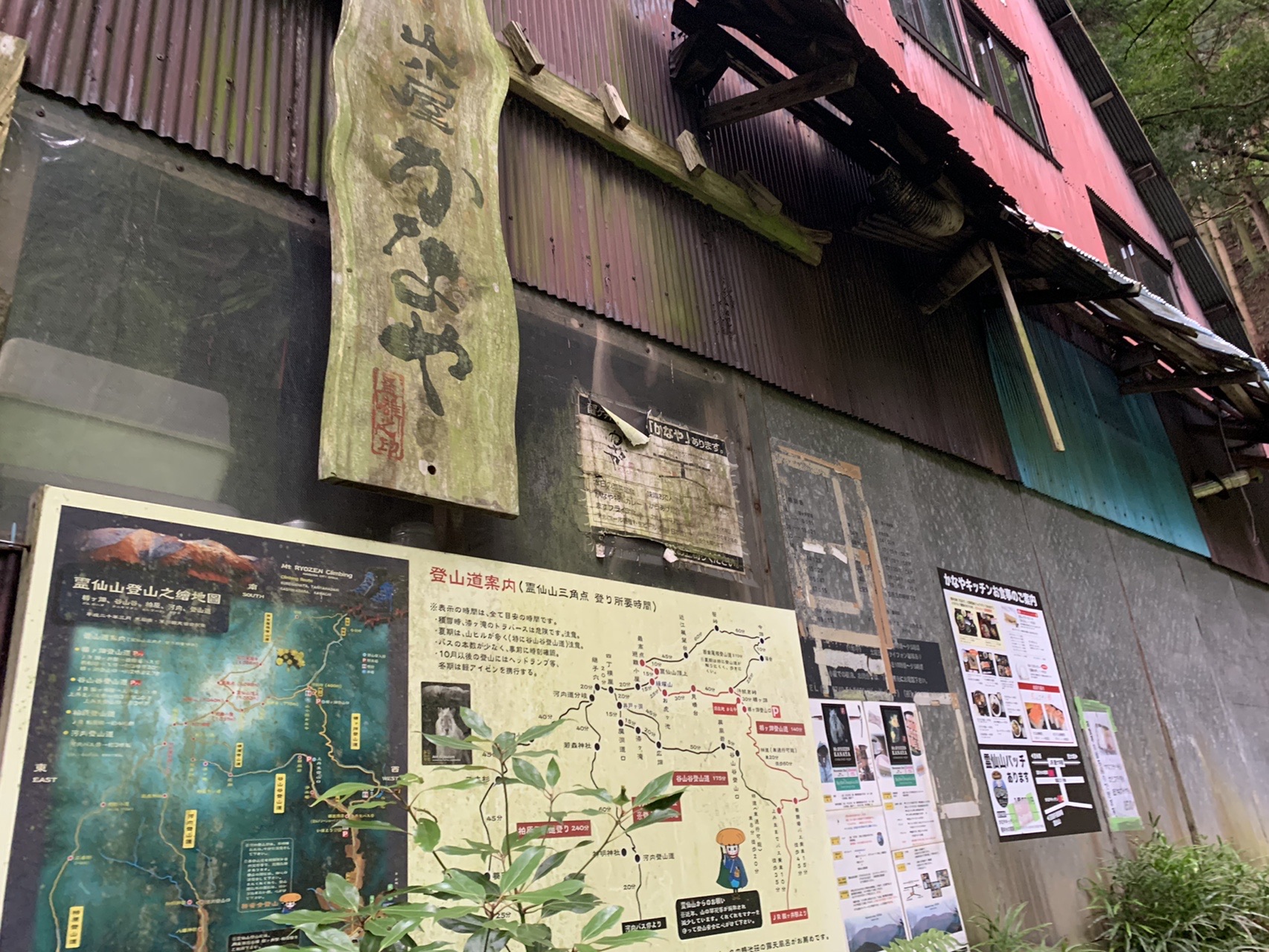 トタン屋根の古い建物の壁に、木の看板と登山マップや掲示物が並んでいる