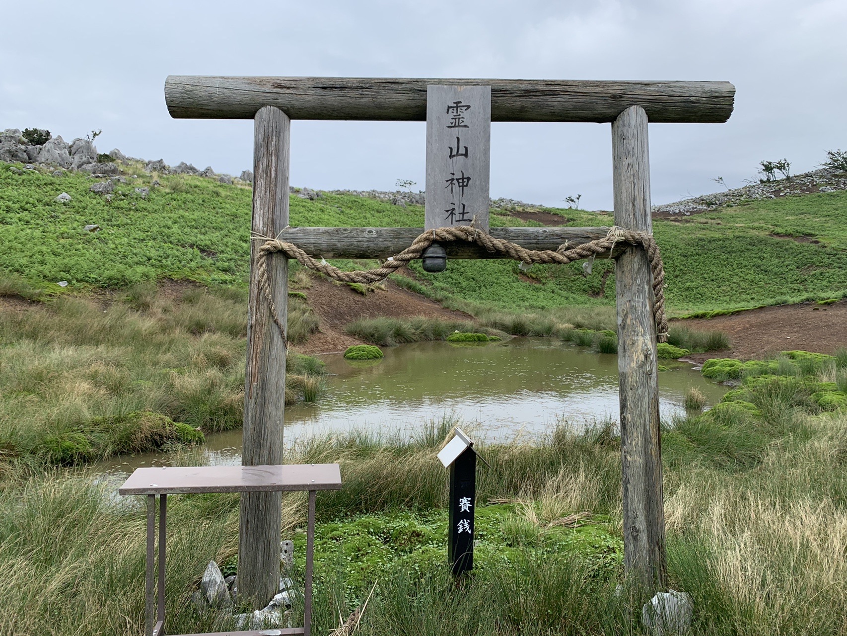 草原の中に立つ木の鳥居と、その奥の小さな池。鳥居には「霊山神社」の文字