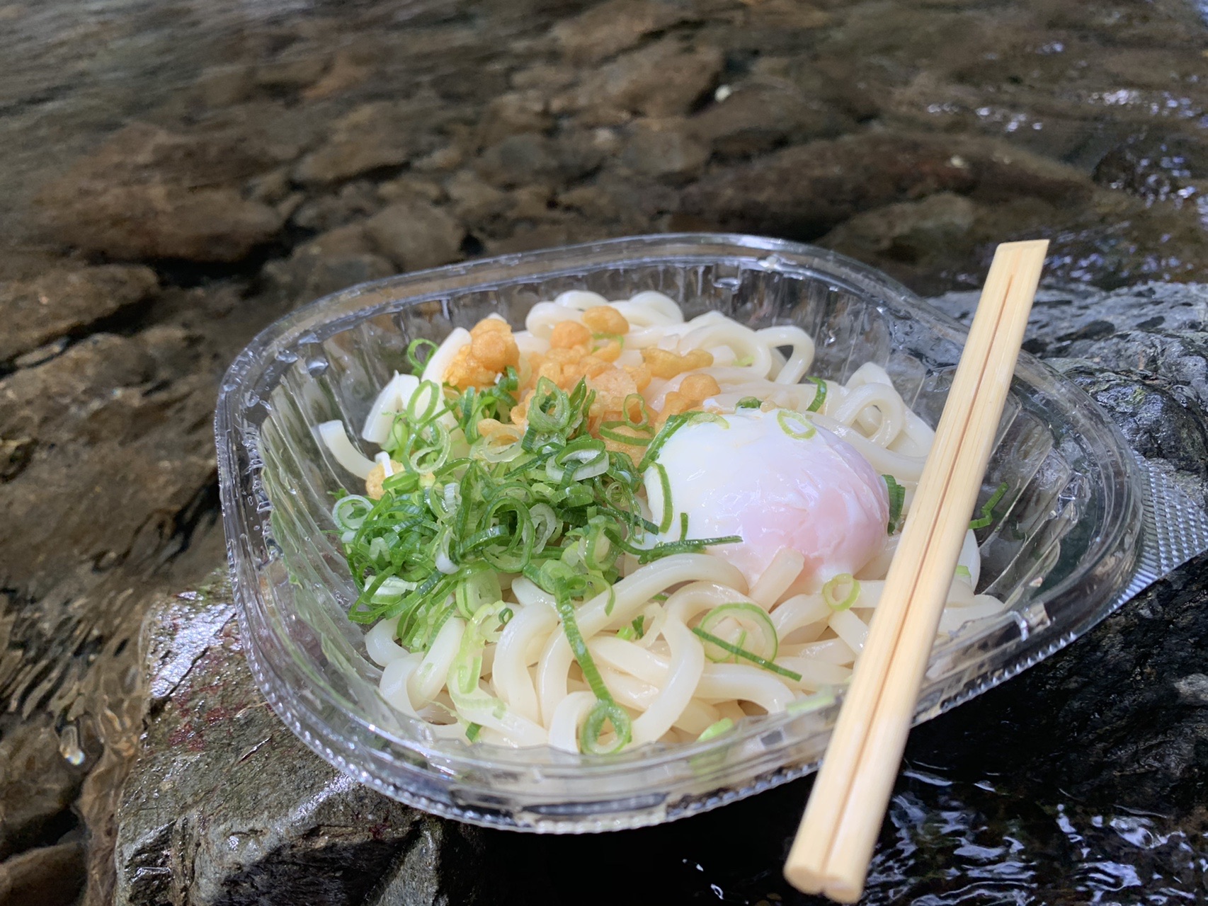 川べりの岩の上に置かれたぶっかけうどんの容器。天かすとネギ、温泉卵がのっている
