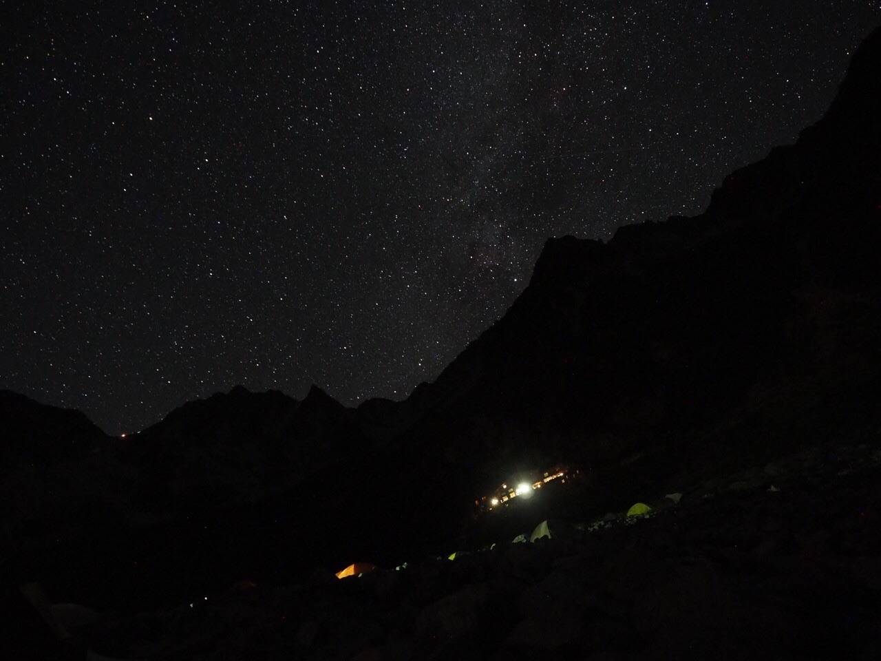 真っ暗な稜線のシルエットの上に無数の星が広がる夜空。ふもとに山小屋の灯り