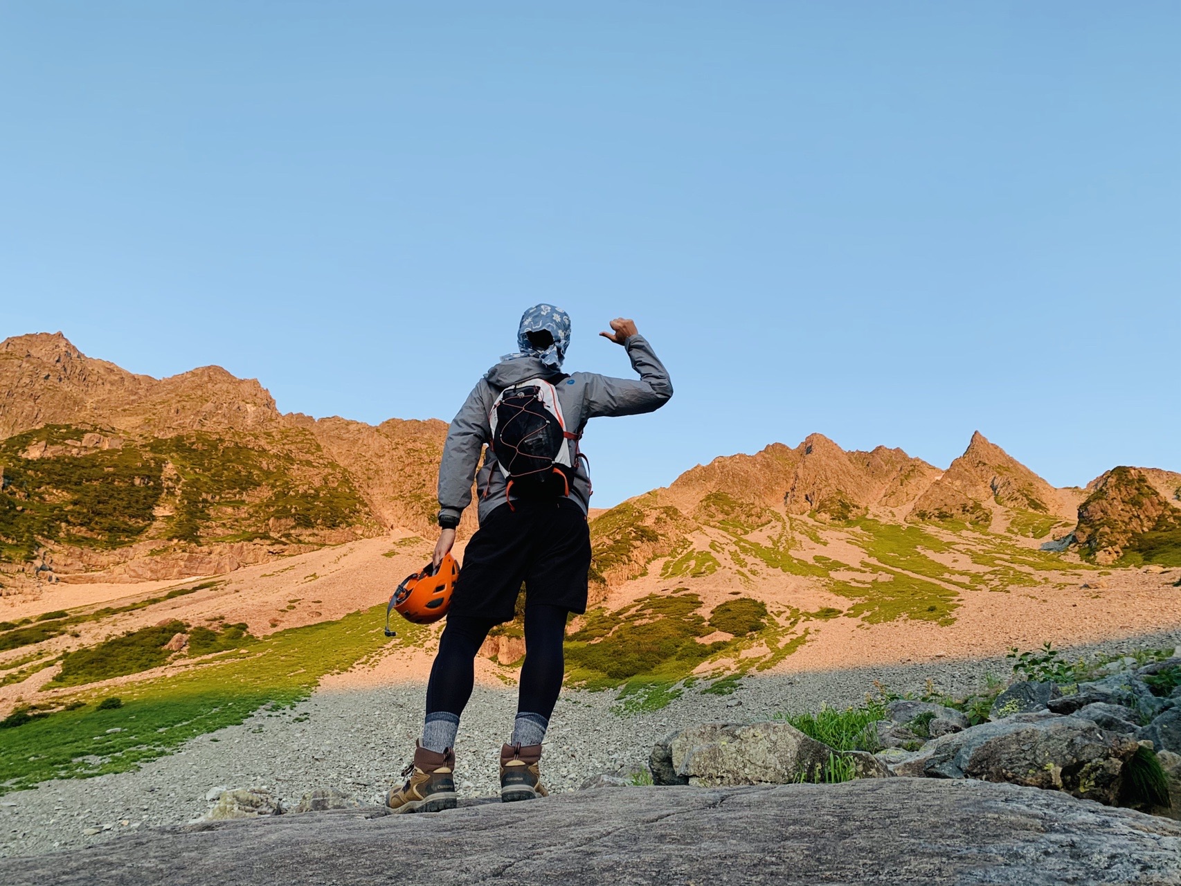 朝焼けに染まる岩壁を背に、ヘルメットをかぶって敬礼するように立つ登山者の後ろ姿
