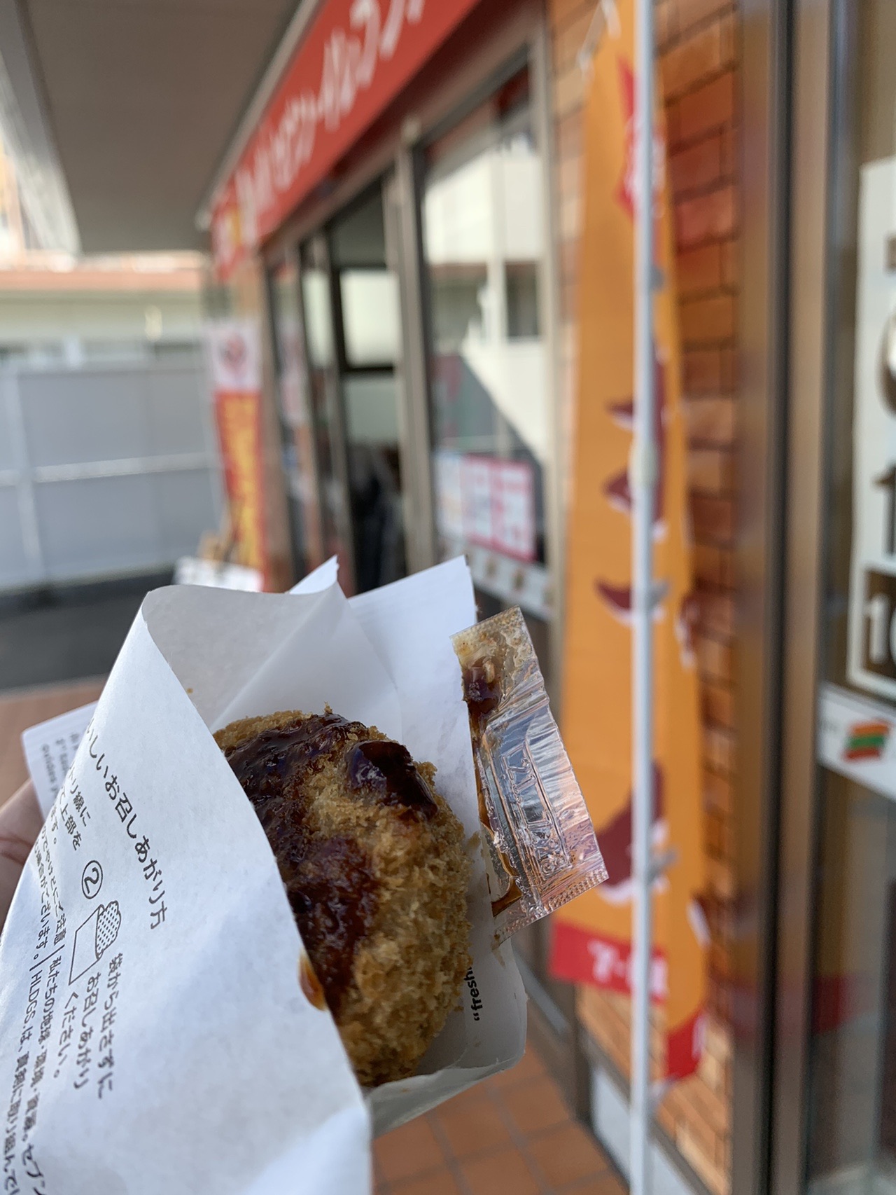 コンビニの店先で紙に包まれたコロッケを持っている手元