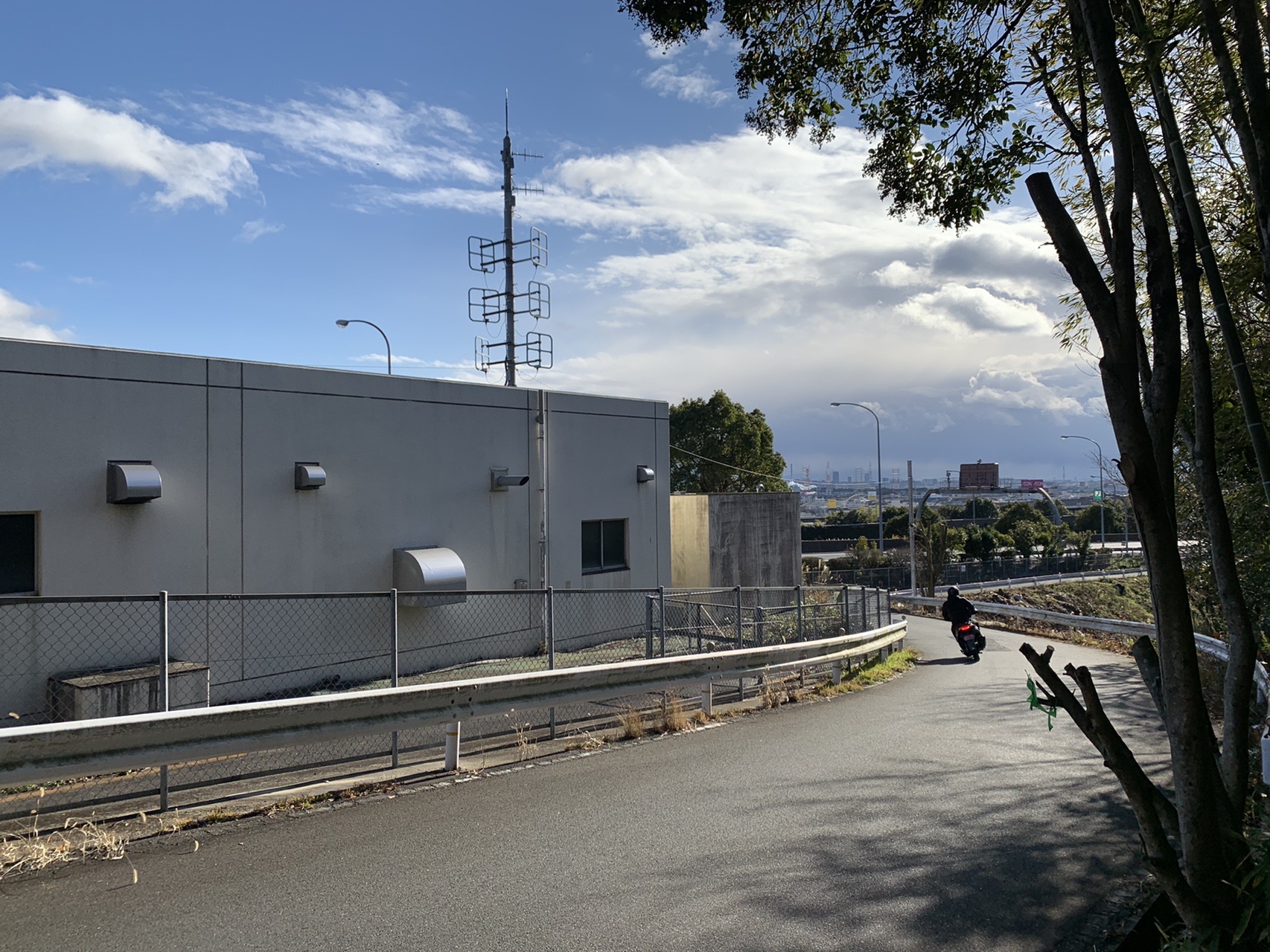 住宅地の脇を抜ける舗装路。奥に鉄塔と街が見える