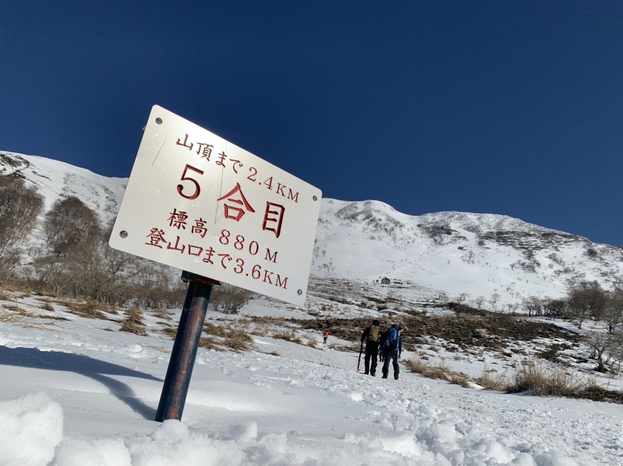 雪原に立つ「5合目」の標識と、遠くを歩く登山者