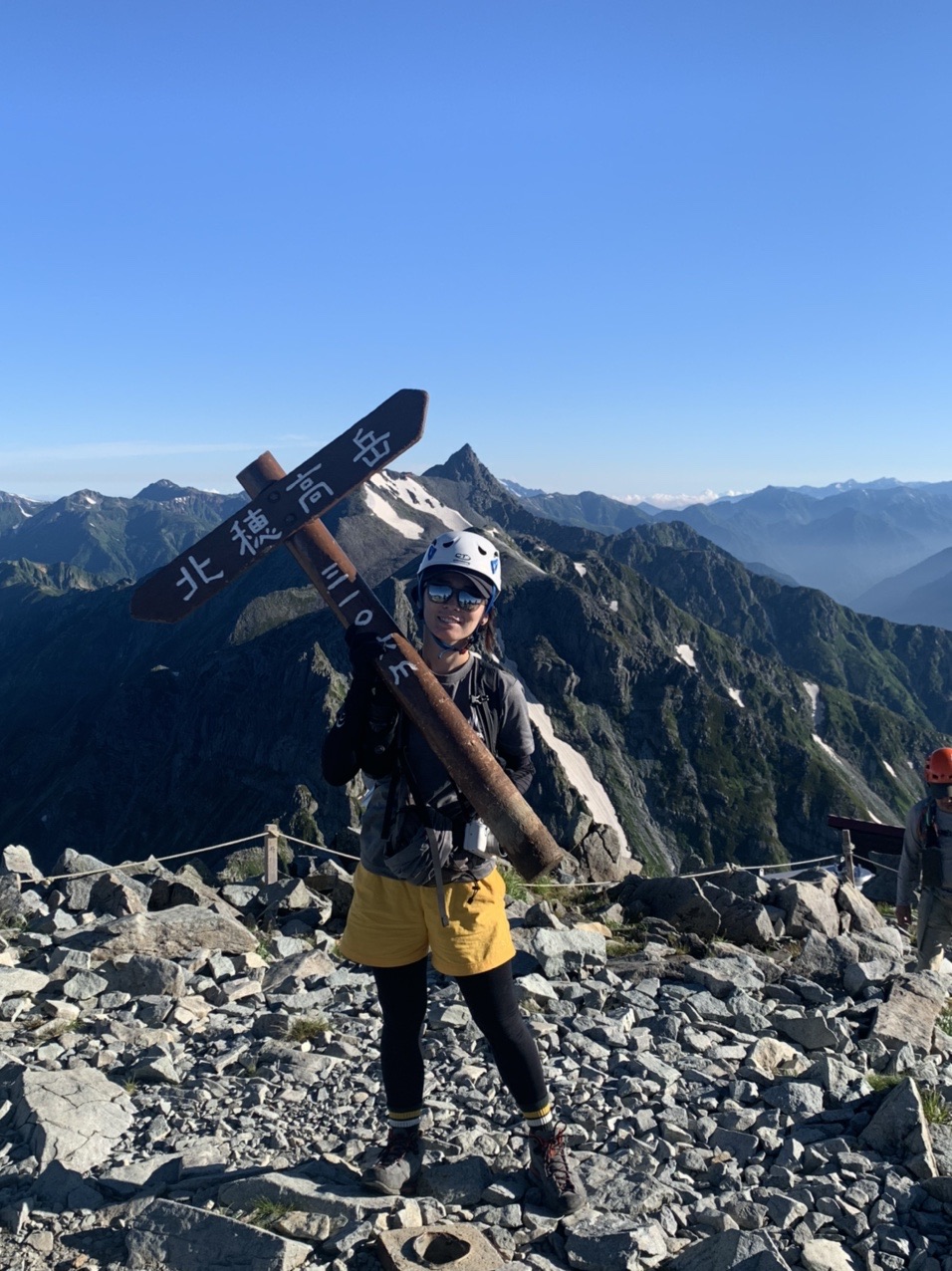 北穂高岳の山頂標識を背に立つ登山者。背後に槍ヶ岳の稜線