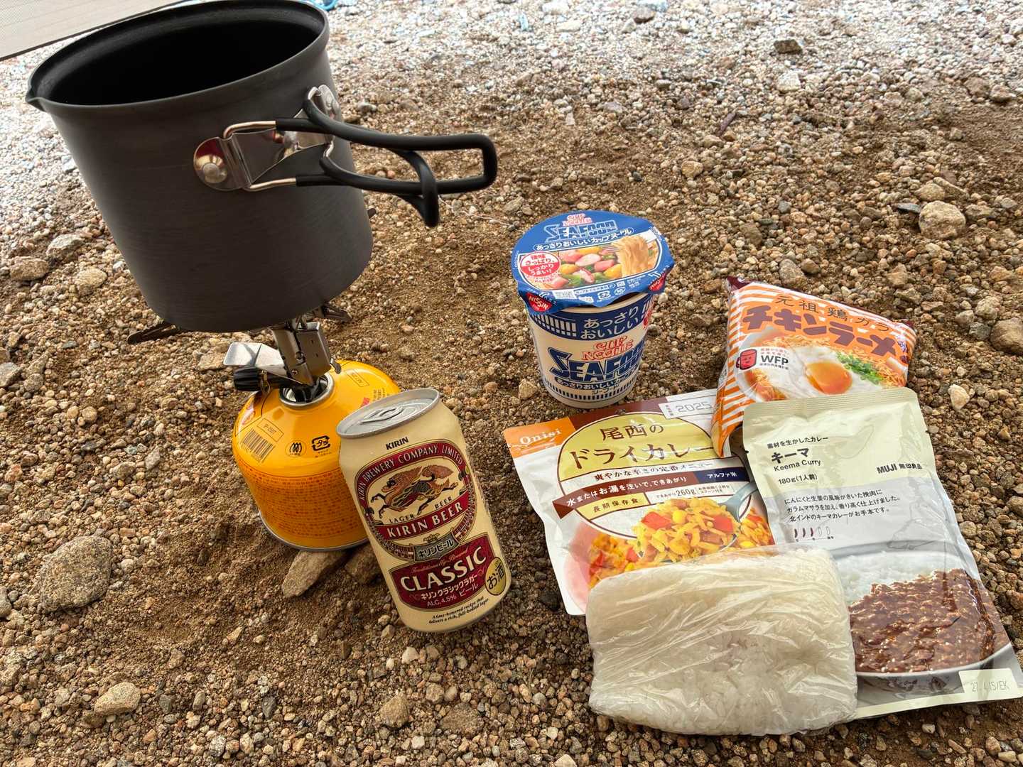 ガスバーナーとクッカー、レトルトのドライカレー、カップ麺、缶ビールが地面に並んでいる