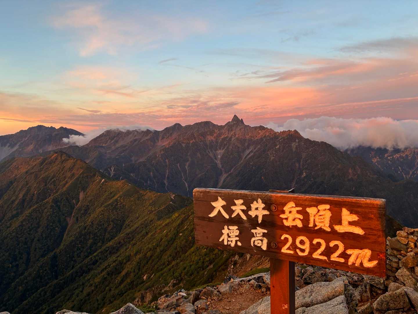 大天井岳の山頂標識「大天井岳頂上 標高2922m」と、背後に朝焼けに染まる槍ヶ岳の稜線